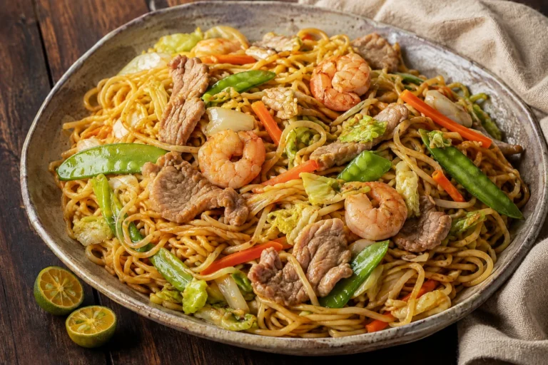 Filipino Pancit Canton with sliced pork, shrimp, carrots, snow peas, cabbage, and calamansi on a ceramic platter.